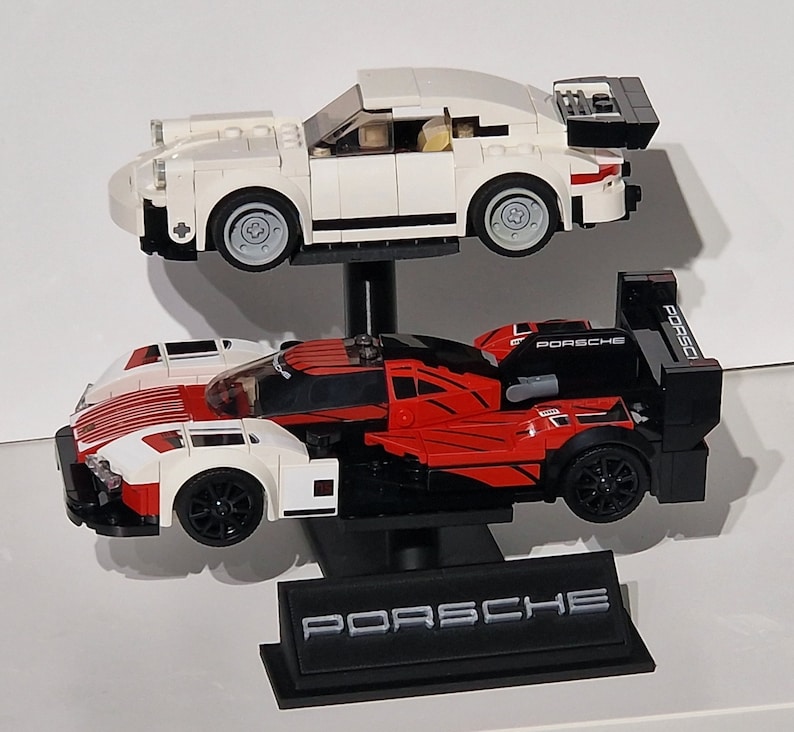 May include: Two toy Porsche race cars, one white and one red and black, are displayed on a black stand. The white car is on top, and the red and black car is below. The stand has the Porsche logo.