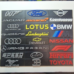 May include: A collection of black rectangular plaques featuring various car brand logos in silver, red, blue, and yellow. Logos include Ford, Jaguar, Lamborghini, Chevrolet, Ferrari, and others. The logos are raised from the surface.