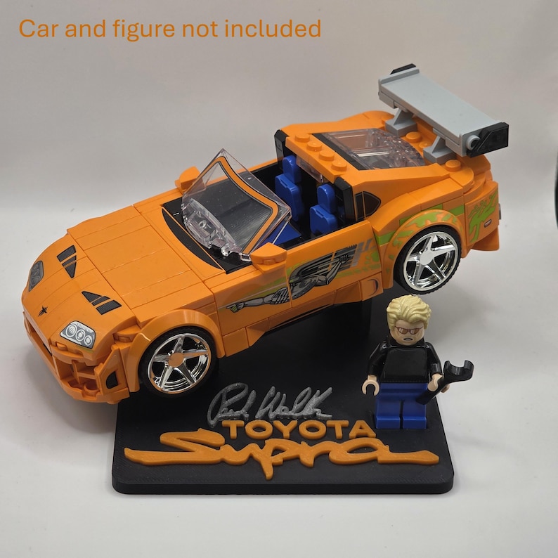 Pu&ograve; includere: Un'auto sportiva giocattolo arancione con alettone posteriore, interni blu e decalcomanie laterali verdi, su un supporto nero con la scritta "Paul Walker TOYOTA Supra" e una piccola statuetta con una chiave inglese. Il testo "Car and figure not included" &egrave; visibile.