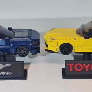 May include: Two toy car models on display stands. A blue Ford Mustang model with a black base and the word "MUSTANG" on the base. A yellow Toyota Supra model with a black base and the word "TOYOTA" on the base.