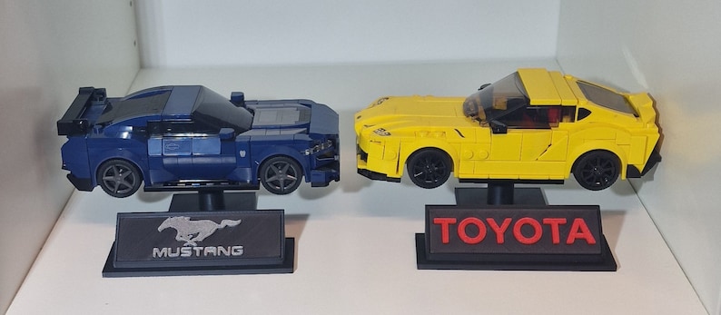 May include: Two toy car models on display stands. A blue Ford Mustang model with a black base and the word "MUSTANG" on the base. A yellow Toyota Supra model with a black base and the word "TOYOTA" on the base.