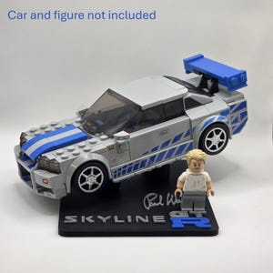 May include: A silver and blue plastic building block sports car model with a rear spoiler, displayed on a black stand with the text SKYLINE GT-R and a signature. The stand includes a small plastic figure. Text states Car and figure not included.