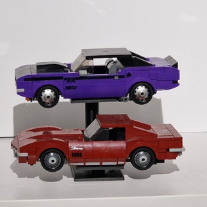 Op de afbeelding: Twee speelgoed-muscle-cars, een paars-zwarte Dodge Challenger en een rode Chevrolet Corvette, worden getoond op een zwarte standaard. De Challenger heeft een zwarte motorkap en de Corvette heeft het woord "Stingray" aan de zijkant.