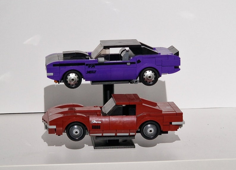 Op de afbeelding: Twee speelgoed-muscle-cars, een paars-zwarte Dodge Challenger en een rode Chevrolet Corvette, worden getoond op een zwarte standaard. De Challenger heeft een zwarte motorkap en de Corvette heeft het woord "Stingray" aan de zijkant.