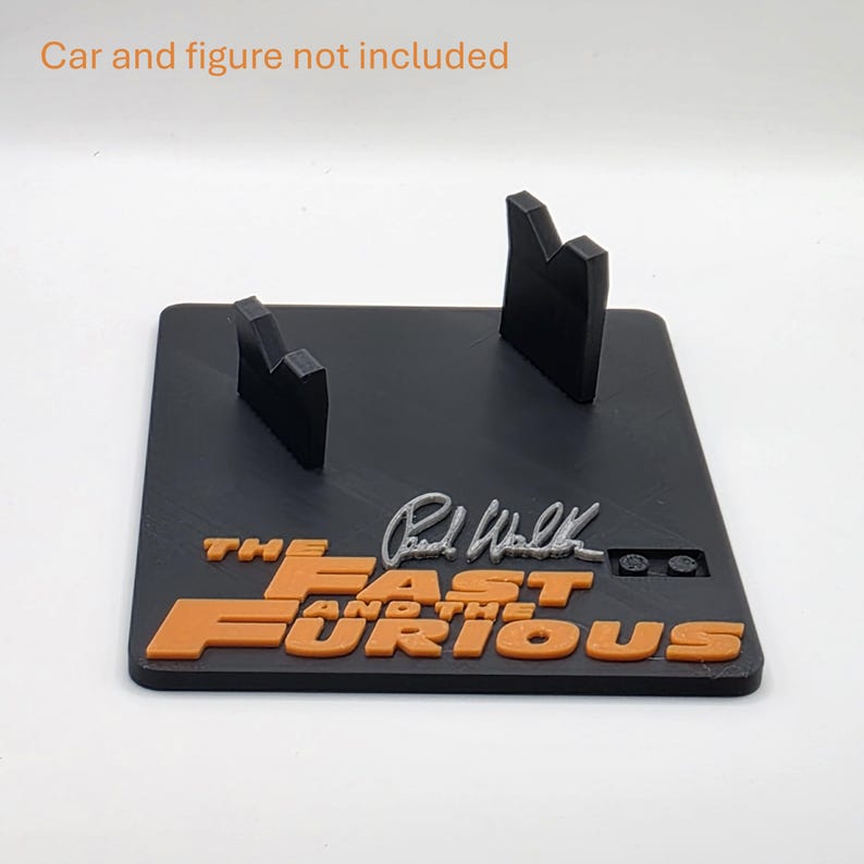 Pu&ograve; includere: Un espositore rettangolare nero con due supporti verticali, il testo arancione in rilievo THE FAST AND THE FURIOUS e una firma argentata. Il testo Car and figure not included &egrave; stampato nella parte superiore dell'immagine.
