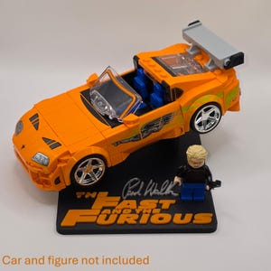 Pu&ograve; includere: Un'auto sportiva giocattolo arancione brillante con un grande alettone posteriore, ruote cromate e decalcomanie laterali verdi, esposta su una base nera con il testo THE FAST AND THE FURIOUS e una firma. Una minifigure bionda si trova accanto all'auto.