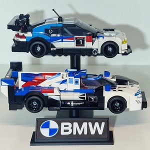 May include: Two Lego race cars, one above the other, in blue, white, and black. The top car has a large spoiler and the number 1. The bottom car has the text "M Motorsport". A black stand with the BMW logo and text supports the cars.
