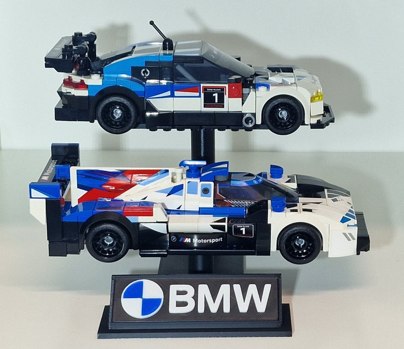 May include: Two Lego race cars, one above the other, in blue, white, and black. The top car has a large spoiler and the number 1. The bottom car has the text "M Motorsport". A black stand with the BMW logo and text supports the cars.