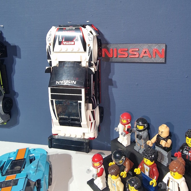 Puede incluir: Un coche de carreras Lego Nissan blanco, negro y rojo con la palabra "NISSAN" impresa en el lateral. El coche est&aacute; montado en una pared con un cartel de pl&aacute;stico rojo que dice "NISSAN". Hay varias minifiguras de Lego en primer plano.