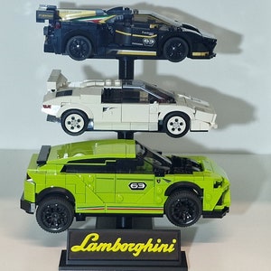 Puede incluir: Un expositor con tres modelos de coches Lamborghini. El coche de arriba es negro con una franja blanca y el texto "Fastron 63". El coche del medio es blanco y el coche de abajo es verde. El expositor tiene una base negra con la palabra "Lamborghini" en letras amarillas.