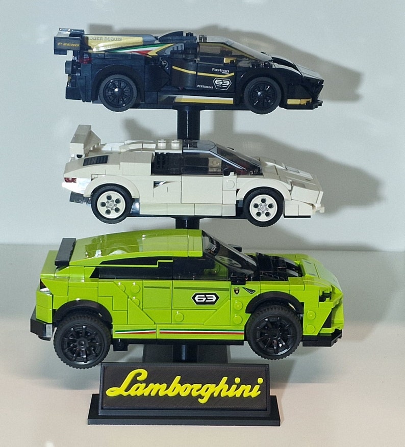 Puede incluir: Un expositor con tres modelos de coches Lamborghini. El coche de arriba es negro con una franja blanca y el texto "Fastron 63". El coche del medio es blanco y el coche de abajo es verde. El expositor tiene una base negra con la palabra "Lamborghini" en letras amarillas.