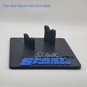 May include: A black display stand featuring two upright supports, a silver Paul Walker signature, and the blue text 2 FAST 2 FURIOUS. The text Car and figure not included is printed at the top. The base is square with a matte finish.