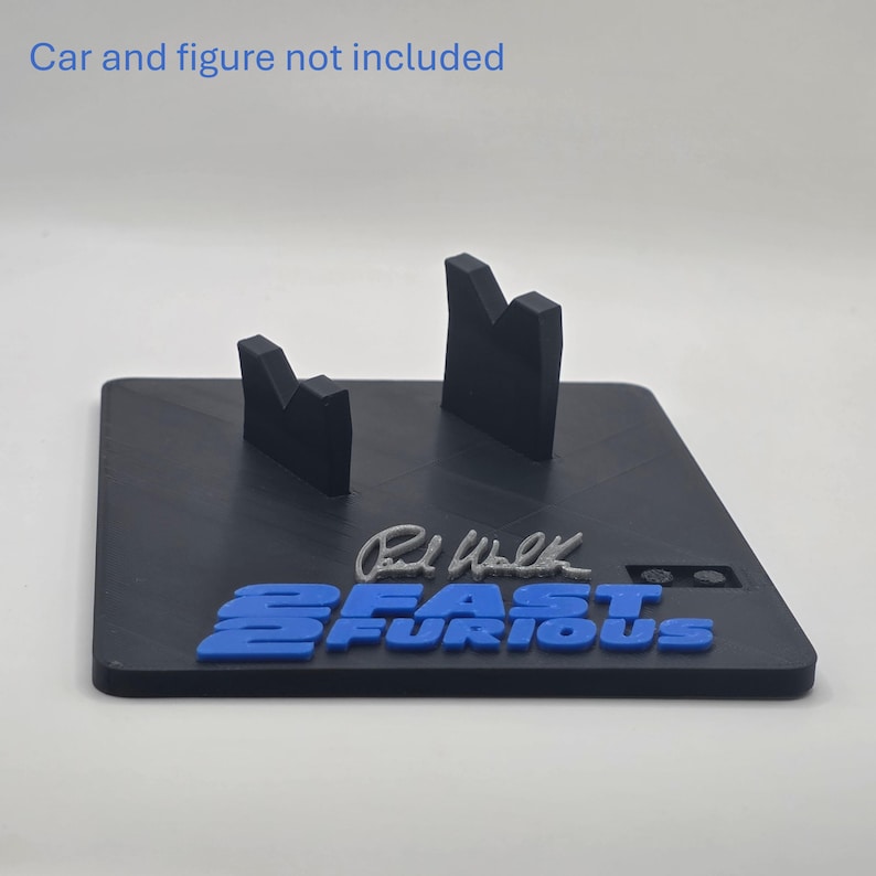 May include: A black display stand featuring two upright supports, a silver Paul Walker signature, and the blue text 2 FAST 2 FURIOUS. The text Car and figure not included is printed at the top. The base is square with a matte finish.