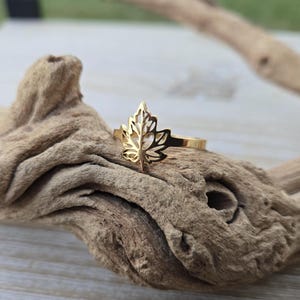 Puede incluir: Un anillo dorado con un diseño detallado de hoja de arce. El anillo se encuentra sobre un fondo de madera natural, resaltando el intrincado patrón recortado de la hoja. Es una pieza de joyería.