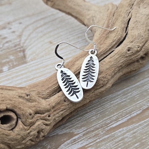 Puede incluir: Pendientes de plata con diseño de árbol. Los pendientes son de forma ovalada y tienen un gancho de plata.