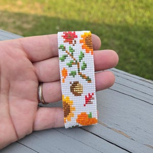 Peut inclure: Marque-page perl&eacute; sur le th&egrave;me de l'automne. Le fond blanc est orn&eacute; de tournesols, de citrouilles et de feuilles aux tons orange, rouge et jaune. Un gland dor&eacute; est &eacute;galement pr&eacute;sent dans le motif.