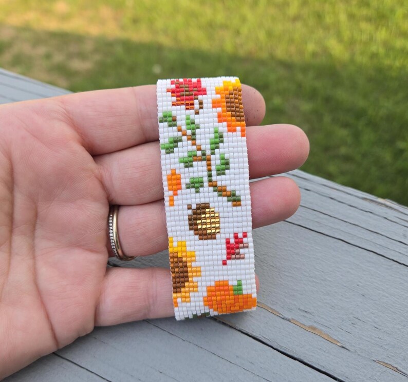 Peut inclure: Marque-page perl&eacute; sur le th&egrave;me de l'automne. Le fond blanc est orn&eacute; de tournesols, de citrouilles et de feuilles aux tons orange, rouge et jaune. Un gland dor&eacute; est &eacute;galement pr&eacute;sent dans le motif.