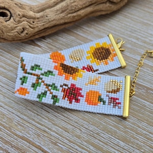 Peut inclure: Deux bracelets perl&eacute;s blancs avec fermoirs et cha&icirc;ne dor&eacute;s. Les bracelets pr&eacute;sentent un motif sur le th&egrave;me de l'automne avec des tournesols, des citrouilles et des feuilles d'automne en orange, marron et rouge. Les bracelets sont sur une surface en bois.