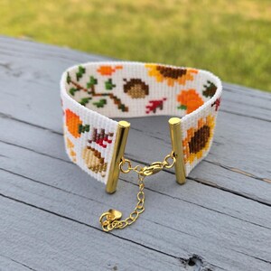 Peut inclure: Un bracelet manchette blanc avec un motif de point de croix sur le th&egrave;me de l'automne. Le motif comprend des tournesols, des citrouilles, des glands et des feuilles d'automne dans des tons orange, marron et vert. Le bracelet a des accents m&eacute;talliques dor&eacute;s et un fermoir &agrave; cha&icirc;ne.