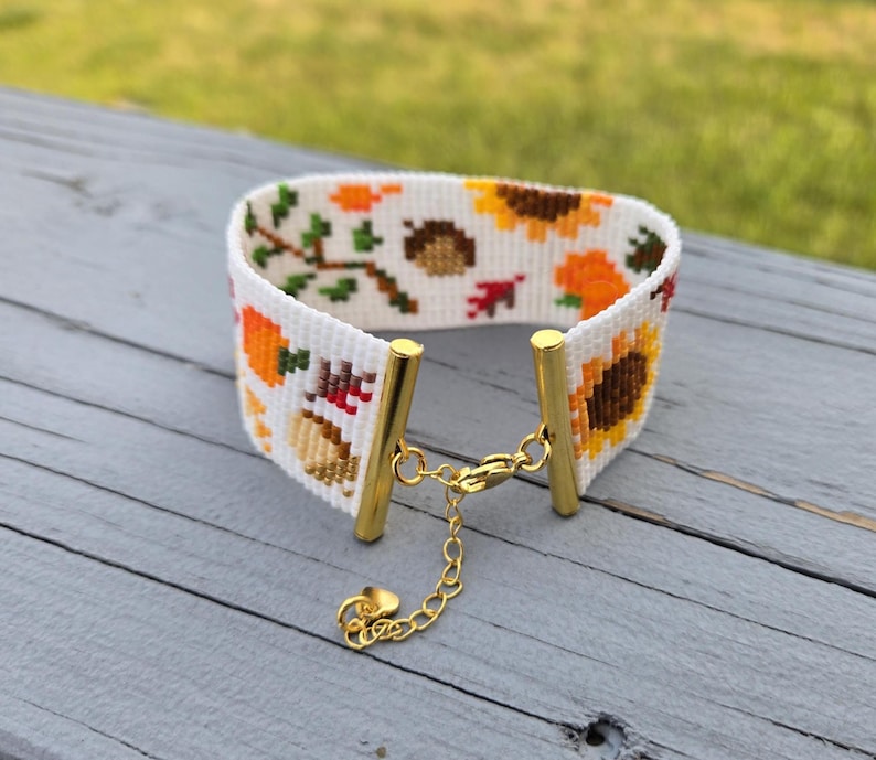 Peut inclure: Un bracelet manchette blanc avec un motif de point de croix sur le th&egrave;me de l'automne. Le motif comprend des tournesols, des citrouilles, des glands et des feuilles d'automne dans des tons orange, marron et vert. Le bracelet a des accents m&eacute;talliques dor&eacute;s et un fermoir &agrave; cha&icirc;ne.
