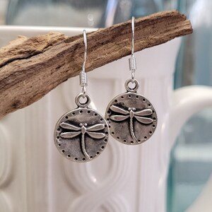 May include: Silver dangle earrings with a dragonfly design. The dragonfly is centered on a round disc with a dotted border.