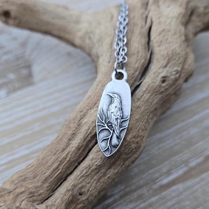 May include: A silver necklace with an oval pendant featuring a detailed, embossed image of a bird perched on a branch.