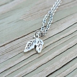 Puede incluir: Un collar de cadena de plata con un delicado colgante que presenta dos hojas con grabado detallado.