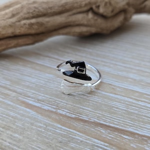 May include: A silver ring with a black witch hat design. The ring has a wrap-around style with a broom detail. The witch hat has a buckle and is painted black. The ring is set against a light wood background.