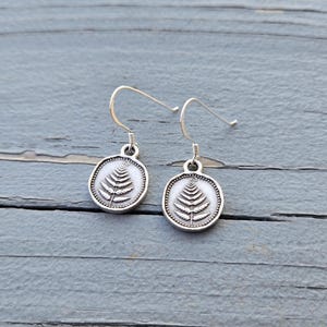 May include: A pair of silver-toned earrings featuring a detailed fern design within a circular frame. The earrings have a hook closure and are displayed on a weathered, gray wooden surface. The fern design is in the center of the earrings.