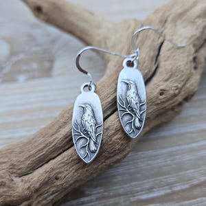 May include: A pair of silver earrings featuring a detailed bird design on each oval-shaped charm. The bird is perched on a branch with intricate details.