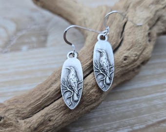 Könnte beinhalten: Ein Paar silberne Ohrringe mit einem detaillierten Vogeldesign auf jedem ovalen Anhänger. Der Vogel sitzt auf einem Ast mit filigranen Details.