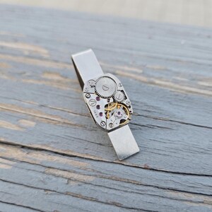 Puede incluir: Un pasador de corbata plateado con un diseño de mecanismo de reloj. El pasador tiene forma rectangular con una exhibición detallada de engranajes, joyas y otros componentes internos. El fondo es una superficie de madera gris desgastada.