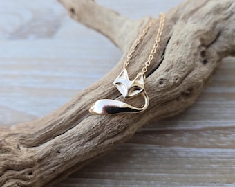 May include: A gold-colored fox pendant necklace. The fox pendant has a detailed head and a curved tail. The necklace is displayed on a piece of driftwood against a light wood background. The chain is delicate and gold.
