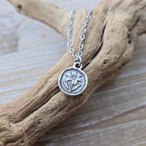 May include: A silver-colored necklace featuring a circular pendant with a detailed lily flower design. The pendant is attached to a delicate chain. The necklace is displayed on a piece of driftwood, creating a natural aesthetic.