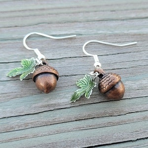 Puede incluir: Un par de pendientes de plata en forma de bellota con detalles de hojas verdes. Las bellotas son de color bronce oscuro.