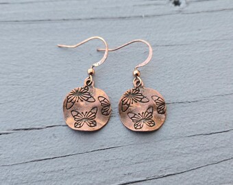 May include: Copper-colored earrings with a butterfly design. Each earring features a circular pendant with etched butterfly patterns. The earrings have a hook closure and are displayed against a blue-gray wooden surface.