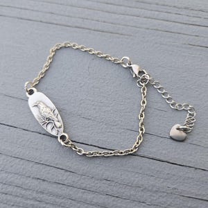 May include: Silver-toned bracelet featuring an oval pendant with a detailed bird design. The bird is perched on a branch, and the bracelet has a delicate chain with an adjustable length and a heart-shaped charm.