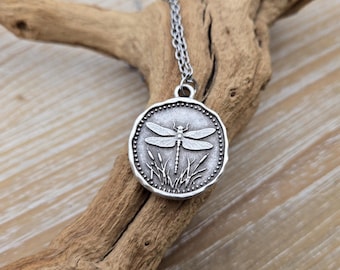 May include: Silver-toned dragonfly pendant necklace. The round pendant features a detailed dragonfly design with reeds, framed by a dotted border. The necklace hangs on a delicate chain, displayed on a piece of driftwood.