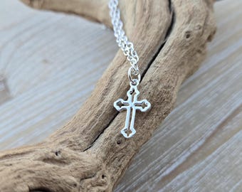 May include: A silver chain necklace with a delicate, openwork cross pendant. The necklace is displayed on a piece of driftwood.
