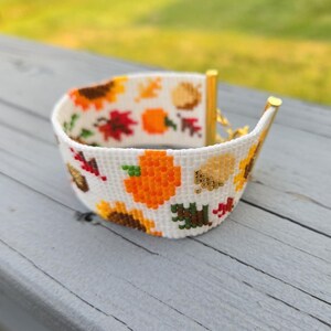 Peut inclure: Un bracelet blanc perl&eacute; sur le th&egrave;me de l'automne. Le bracelet pr&eacute;sente un motif de tournesols, de citrouilles et de feuilles d'automne en orange, jaune, rouge et vert. Le bracelet a des extr&eacute;mit&eacute;s en m&eacute;tal dor&eacute;.