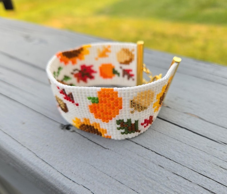Peut inclure: Un bracelet blanc perl&eacute; sur le th&egrave;me de l'automne. Le bracelet pr&eacute;sente un motif de tournesols, de citrouilles et de feuilles d'automne en orange, jaune, rouge et vert. Le bracelet a des extr&eacute;mit&eacute;s en m&eacute;tal dor&eacute;.