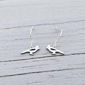 Op de afbeelding: Een paar zilveren oorhangers met een vogel-silhouetontwerp. De oorbellen zijn gemaakt van metaal en hebben een eenvoudige, elegante stijl.
