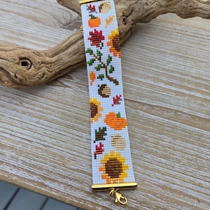 Peut inclure: Un bracelet blanc perl&eacute; sur le th&egrave;me de l'automne. Le bracelet pr&eacute;sente des tournesols, des citrouilles et des feuilles en orange, jaune, marron et vert. Le bracelet a des extr&eacute;mit&eacute;s en m&eacute;tal dor&eacute; et un fermoir.