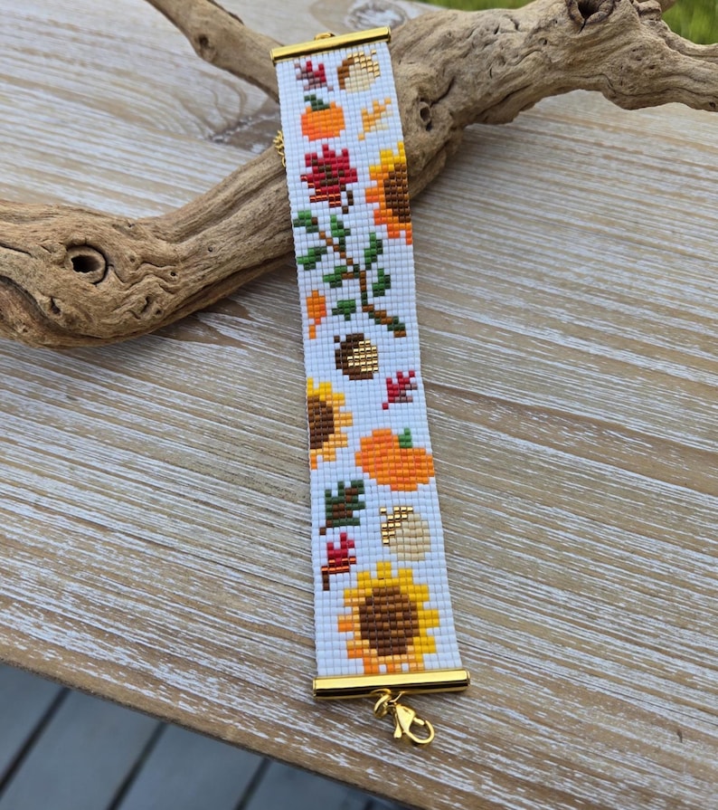 Peut inclure: Un bracelet blanc perl&eacute; sur le th&egrave;me de l'automne. Le bracelet pr&eacute;sente des tournesols, des citrouilles et des feuilles en orange, jaune, marron et vert. Le bracelet a des extr&eacute;mit&eacute;s en m&eacute;tal dor&eacute; et un fermoir.