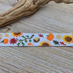 Peut inclure: Bracelet de perles avec un motif d'automne. Il pr&eacute;sente des tournesols, des citrouilles et des feuilles aux couleurs orange, jaune et rouge. Le bracelet a des fermoirs dor&eacute;s et un fond bleu clair. Le design comprend des glands.