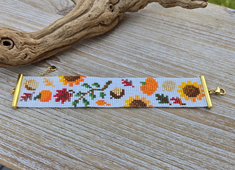 Peut inclure: Bracelet de perles avec un motif d'automne. Il pr&eacute;sente des tournesols, des citrouilles et des feuilles aux couleurs orange, jaune et rouge. Le bracelet a des fermoirs dor&eacute;s et un fond bleu clair. Le design comprend des glands.