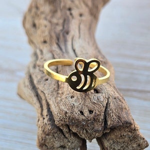 May include: A gold-colored ring featuring a bee design. The ring has an adjustable band and a cutout bee silhouette. The ring is displayed on a piece of driftwood, showcasing its details and design.
