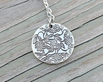 Puede incluir: Un collar de plata con un colgante redondo que presenta un delicado diseño floral. El colgante está grabado con un patrón de flores y hojas.