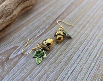 Puede incluir: Un par de pendientes de bellota dorados con detalles de hojas verdes. Los pendientes son de estilo colgante con cierre de gancho.