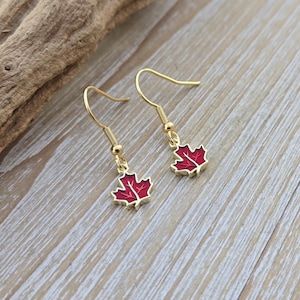 May include: A pair of gold dangle earrings with red maple leaf charms. The earrings are on a wooden surface.